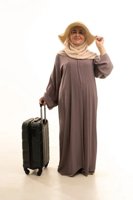 مفهوم السياحة فى إجازة الصيف, الاستعداد لقضاء رحلة صيفية ممتعة , النظر الى الكاميرا بايماءات السعادة و السرور، بورتريه لامرأة عربية خليجية سعودية كبيرة في السن ترتدي العباءة و الحجاب مع قبعة من القش تمسك بيدها حقيبة سفر , صورة للجسم بالطول الكامل , خلفية بيضاء