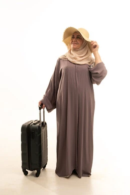 Looking at something with gestures of joy and happiness, preparing for travel and trips, going on an enjoyable summer vacation, a portrait of an elderly Saudi Arabian Gulf woman wearing an abaya and hijab with a straw hat holding a travel bag in her hand, a full-body image, white background.