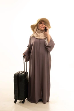 اجراء مكالمة هاتفية عبرالجوال، النظر بإيماءات الفرح و السرور، التجهيز للسفر و الرحلات، الذهاب في عطلة صيفية ممتعة  , بورتريه لامرأة عربية خليجية سعودية كبيرة في السن ترتدي العباءة و الحجاب مع قبعة من القش تمسك بيدها حقيبة سفر , صورة للجسم بالطول الكامل , خلفية بيضاء