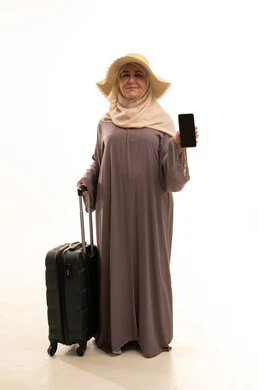 A blank black screen displayed on a mobile phone, looking with gestures of joy and happiness, preparing for travel and trips, going on a fun summer vacation, a portrait of an elderly Saudi Arabian Gulf woman wearing an abaya and hijab with a straw hat holding a travel bag in her hand, a full-body image, white background.