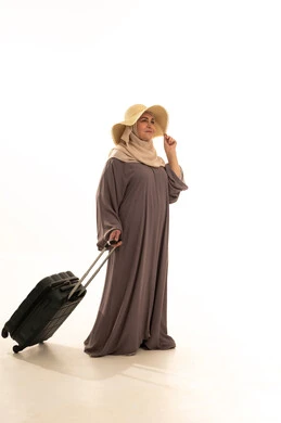 النظر بإيماءات الفرح و السرور، التجهيز للسفر و الرحلات، الذهاب في عطلة صيفية ممتعة  , بورتريه لامرأة عربية خليجية سعودية كبيرة في السن ترتدي العباءة و الحجاب مع قبعة من القش تمسك بيدها حقيبة سفر , صورة للجسم بالطول الكامل , خلفية بيضاء