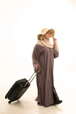 الذهاب في عطلة صيفية ممتعة , التجهيز للسفر و الرحلات , بورتريه لامرأة عربية خليجية سعودية كبيرة في السن ترتدي العباءة و الحجاب مع قبعة من القش تمسك بيدها حقيبة سفر , النظر بإيماءات الفرح و السرور , صورة للجسم بالطول الكامل , خلفية بيضاء