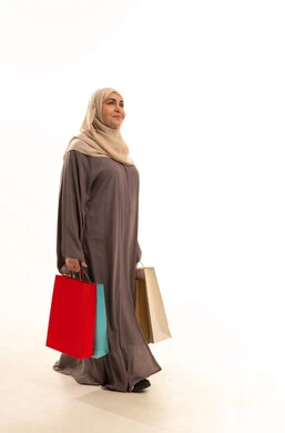 Annual offers and discounts in the markets, shopping and buying supplies and gifts, a portrait of an elderly Saudi Gulf Arab woman wearing an abaya and hijab carrying shopping bags in her hands, gestures indicating movement and walking, a full-body image, white background.
