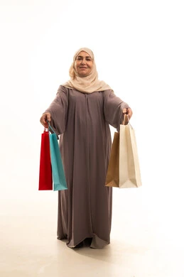 Shopping and buying supplies and gifts, getting offers and discounts, a portrait of an elderly Saudi Gulf Arab woman wearing an abaya and hijab holding shopping bags in her hands, looking at the camera with gestures of joy and happiness, a full-body image, white background.