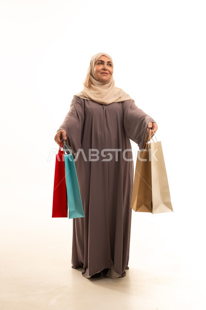 Getting offers and discounts, shopping and buying supplies and gifts, a portrait of an elderly Saudi Gulf Arab woman wearing an abaya and hijab holding shopping bags in her hands, looking at something with gestures of joy and happiness, a full-body image, white background.