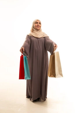 Getting offers and discounts, shopping and buying supplies and gifts, a portrait of an elderly Saudi Gulf Arab woman wearing an abaya and hijab holding shopping bags in her hands, looking at something with gestures of joy and happiness, a full-body image, white background.