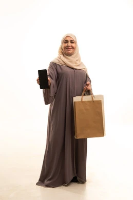 التسوق و شراء المستلزمات المنزلية , استخدام الأجهزة التقنية الحديثة , بورتريه لامرأة عربية خليجية سعودية كبيرة في السن ترتدي الثوب التقليدي و الحجاب تحمل بيدها أكياس المشتريات و الهاتف المحمول بشاشة سوداء فارغة , صورة للجسم بالطول الكامل , خلفية بيضاء