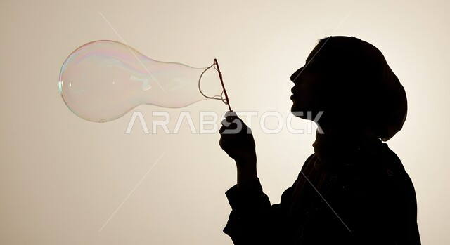 الاستمتاع بصنع فقاعة الصابون في الهواء, قضاء الأوقات بالأنشطة الترفيهية الممتعه والمسلية, اللعب بالفقاعات المائية المتطايرة والمتناثرة، صورة ظلية لامرأة عربية خليجية سعودية تقوم بنفخ فقاعة صابون كبيرة