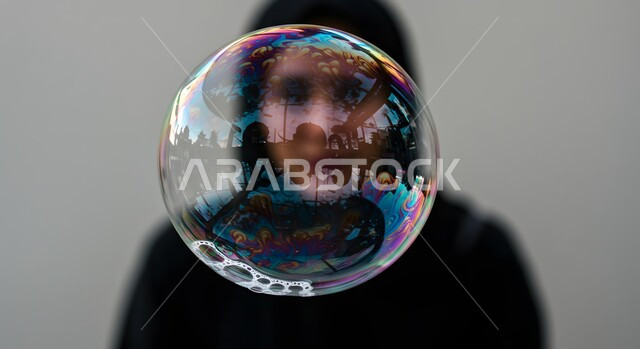 Entertainment and recreation with children's games, enjoying making soap bubbles in the air, spending time with fun and entertaining recreational activities, playing with flying and scattering water bubbles, a close-up image of a large soap bubble reflected on a Saudi Arabian Gulf woman