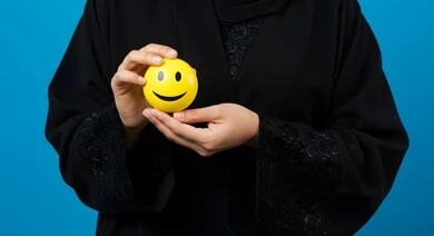 Emoticons and expressions, portrait of a Saudi Arabian Gulf woman holding a yellow smiley face, happy face emoji, positive thinking concept, excellent service rating, blue background