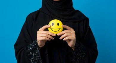 Emoticons and expressions, portrait of a Saudi Arabian Gulf woman holding a yellow smiley face, happy face emoji, positive thinking concept, excellent service rating, blue background