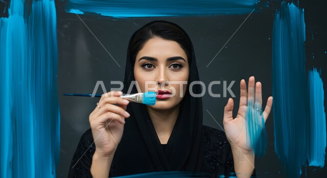 نشاطات ترفيهية فنية، قضاء اوقات ممتعة في الرسم, صورة مقربة لامرأة عربية خليجية سعودية محجبة تحمل بيدها فرشاة ألوان باللون الأزرق, ممارسة هواية الرسم والتلوين، خلفية سوداء