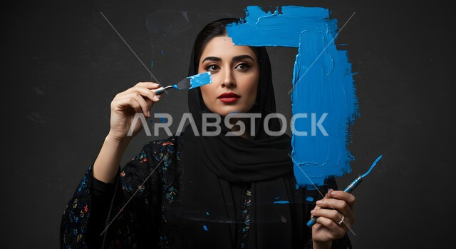 نشاطات ترفيهية فنية، قضاء اوقات ممتعة في الرسم, صورة مقربة لامرأة عربية خليجية سعودية محجبة تحمل بيدها فرشاة ألوان باللون الأزرق, ممارسة هواية الرسم والتلوين، خلفية سوداء