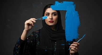 Artistic recreational activities, having fun drawing, close-up of a veiled Saudi Arabian Gulf woman holding a blue paintbrush, practicing the hobby of drawing and coloring, black background
