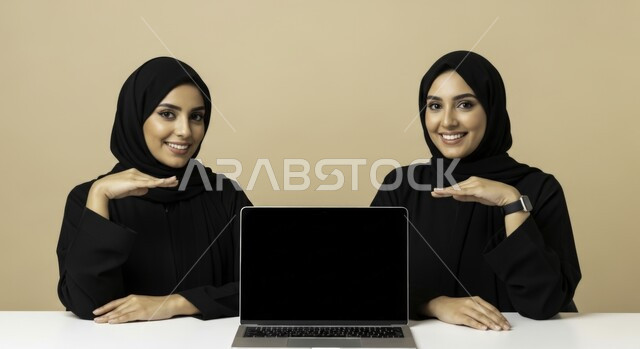 النظر إلى الكاميرا بإيماءات السرور، مهنة ووظيفة نسائية مكتبية سعودية، ادارة وانجاز المهام باستخدام جهاز تقني حديث ومتطور, امرأتان عربيتان خليجيتان سعوديتان محجبتان ترتديان عباءة سوداء تجلسان في المكتب في مقر العمل