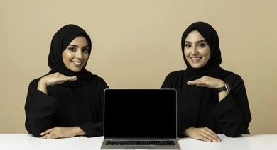 Looking at the camera with gestures of pleasure, a Saudi female office profession and job, managing and completing tasks using a modern and advanced technical device, two veiled Saudi Arabian Gulf women wearing black abaya sitting in the office at the workplace