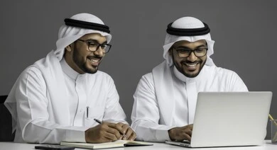 كتابة التقارير والمستندات، التفاهم والتعاون بين الزملاء لتحقيق أهداف الشركة , إدارة شؤون الأعمال وتنظيمها باستخدام الجهاز التقني الحديث، رجلان عربيان خليجيان اماراتيان يرتديان الكندورة والغتره والنظارة الطبية يقومان بالعمل على الحاسوب المحمول داخل المكتب
