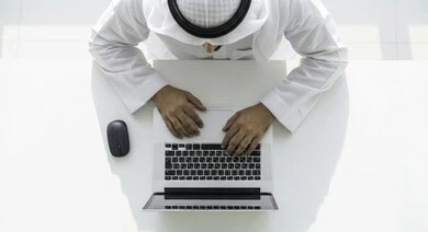 Managing daily work tasks at the company's headquarters, Emirati professions and jobs, using modern technical devices, an overhead image of an Emirati Gulf Arab man wearing a kandura and ghutra sitting in his office working on the computer, developing work plans and mechanisms, integrating work with technology and technology.