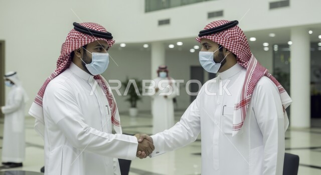 Wearing a medical mask to prevent the Corona virus, taking preventive and precautionary measures, making friends and building social relationships, partnerships in business and projects, two Saudi Gulf Arab men wearing a thobe and a shemagh shaking hands, expanding areas of work