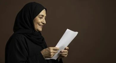 Completing the required tasks, following up on correspondence and records, a smiling elderly Saudi Arabian Gulf woman wearing an abaya and hijab holding white papers in her hands and looking at them, carefully reading notes and reports