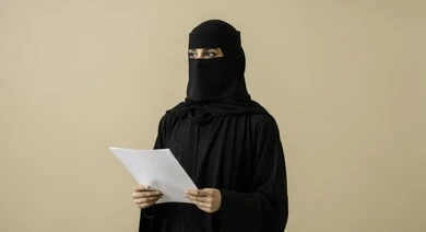 Completing required tasks, following up on correspondence and records, a Saudi Arabian Gulf woman wearing a niqab and abaya holding blank papers in her hands, carefully reading notes and reports, looking at something, following up on plans and reports