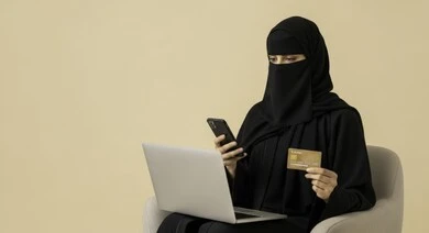 إمكانية الشراء والدفع من خلال البرامج والتطبيقات الالكترونية، استخدام أجهزة تقنية حديثة للدفع عن بعد عبر الانترنت، سيدة عربية خليجية سعودية منقبة ترتدي عباءة تجلس على كرسي وتحمل الهاتف والحاسوب المحمول وبطاقة الإئتمان البنكية للتسوق الالكتروني