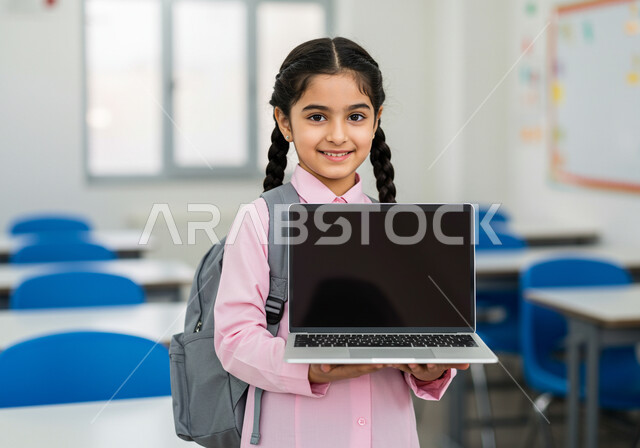 دمج التكنولوجيا بالتعليم في المملكة العربية السعودية, عرض شاشة سوداء فارغة، الاستعداد للعام الدراسي الجديد، تلميذة عربية خليجية سعودية ترتدي اليونيفوورم المدرسي وحقيبة الظهر المدرسية تحمل بيدها حاسوب محمول، إيماءات الحماس والسعادة بالعودة إلى المدرسة