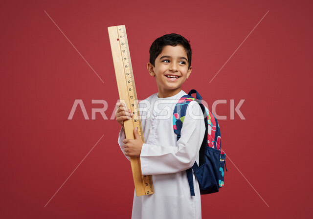العودة إلى المدارس، طالب عربي خليجي سعودي مبتسم يرتدي الزي التقليدي يحمل حقيبة الظهر المدرسية وبيده مسطرة مرقمة يقوم بالنظر الى شيء ما بتعابير السرور، أدوات قرطاسية جديدة، التعليم الدراسي في المملكة العربية السعودية