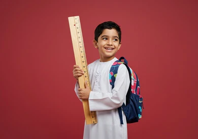 العودة إلى المدارس، طالب عربي خليجي سعودي مبتسم يرتدي الزي التقليدي يحمل حقيبة الظهر المدرسية وبيده مسطرة مرقمة يقوم بالنظر الى شيء ما بتعابير السرور، أدوات قرطاسية جديدة، التعليم الدراسي في المملكة العربية السعودية