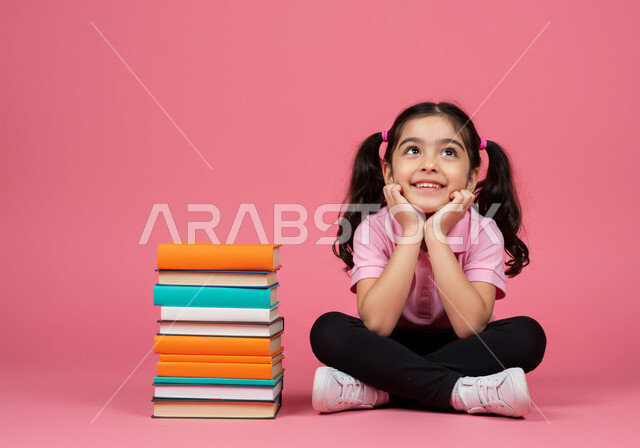 Expressions of happiness and excitement about returning to school in the Kingdom of Saudi Arabia. A portrait of a Saudi Arabian Gulf Arab student sitting on the floor with a group of school books next to her, looking at something with gestures of cheerfulness and pleasure.
