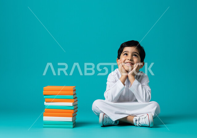 الاستعداد للعام الدراسي الجديد، تعابير السعادة بالعودة للمدارس في المملكة العربية السعودية، بورتريه لطالب عربي خليجي سعودي يرتدي الثوب التقليدي يجلس على الأرض وبجانبه مجموعة من الكتب المدرسية، النظر إلى شيء ما بإيماءات البشاشة والسرور