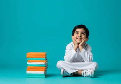 الاستعداد للعام الدراسي الجديد، تعابير السعادة بالعودة للمدارس في المملكة العربية السعودية، بورتريه لطالب عربي خليجي سعودي يرتدي الثوب التقليدي يجلس على الأرض وبجانبه مجموعة من الكتب المدرسية، النظر إلى شيء ما بإيماءات البشاشة والسرور