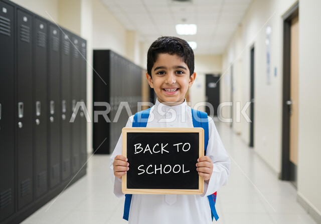 طالب عربي خليجي سعودي يرتدي الزي المدرسي ويحمل حقيبة الظهر وسبورة سوداء صغيرة مكتوب عليها نص العودة إلى المدارس باللغة الانجليزية، جودة التعليم في السعودية، استقبال العام الدراسي الجديد بتعابير السعادة والسرور
