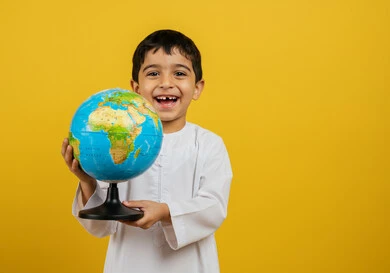 The concept of searching and exploring around the world, a Saudi Arabian Gulf boy wearing traditional clothes holding a globe, looking at the camera with expressions of happiness and pleasure, a spherical world map