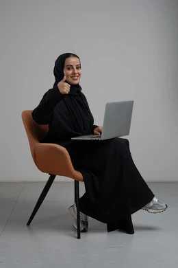 Expressions of approval and acceptance, gestures of satisfaction and admiration, office profession and job, management and completion of tasks using a modern and advanced technical device, portrait of a Saudi Arabian Gulf woman wearing a hijab and a black abaya sitting on a chair and working on a laptop, gray background