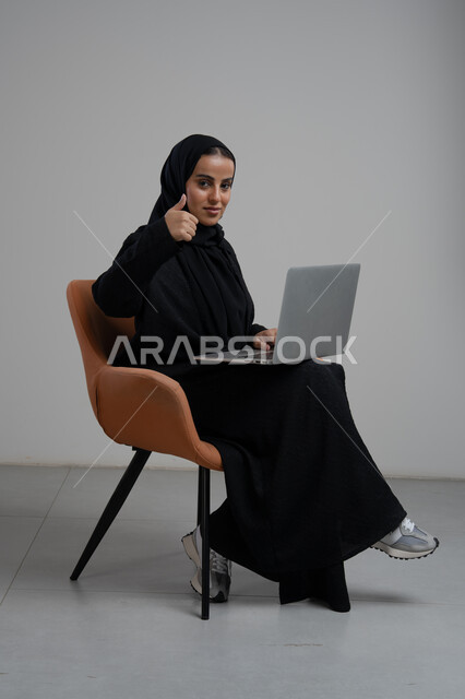 تعابير الموافقة والقبول, ايماءات الرضى والاعجاب، مهنة و وظيفة مكتبية، ادارة وانجاز المهام باستخدام جهاز تقني حديث ومتطور, بورتريه لإمرأة عربية خليجية سعودية ترتدي الحجاب والعباءة السوداء تجلس على الكرسي وتعمل على الحاسوب المحمول, خلفية رمادية