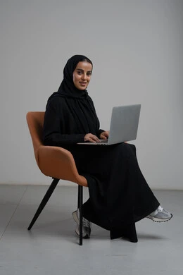 Office profession and job, managing and completing tasks using a modern and advanced technical device, portrait of a Saudi Arabian Gulf woman wearing a hijab and a black abaya sitting on a chair and working on a laptop, gray background