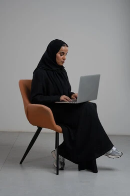 Office profession and job, managing and completing tasks using a modern and advanced technical device, portrait of a Saudi Arabian Gulf woman wearing a hijab and a black abaya sitting on a chair and working on a laptop, gray background