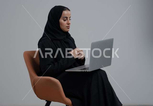 مهنة و وظيفة مكتبية سعودية، ادارة وانجاز المهام باستخدام جهاز تقني حديث ومتطور, بورتريه لإمرأة عربية خليجية سعودية ترتدي الحجاب والعباءة السوداء تجلس على الكرسي و تعمل على الحاسوب المحمول, خلفية رمادية
