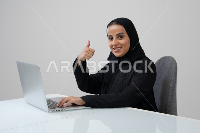 رفع اصبع الابهام بايماءات الرضى، تعابير الموافقة والقبول, مهنة ووظيفة مكتبية، ادارة وانجاز المهام باستخدام جهاز تقني حديث ومتطور, بورتريه لإمرأة عربية خليجية سعودية ترتدي الحجاب والعباءة السوداء تعمل على الحاسوب المحمول في مقر الشركة, خلفية رمادية