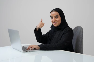 Thumbs up gesture, expressions of approval and acceptance, office profession and job, managing and completing tasks using a modern and advanced technical device, portrait of a Saudi Arabian Gulf woman wearing a hijab and black abaya working on a laptop at the company headquarters, gray background