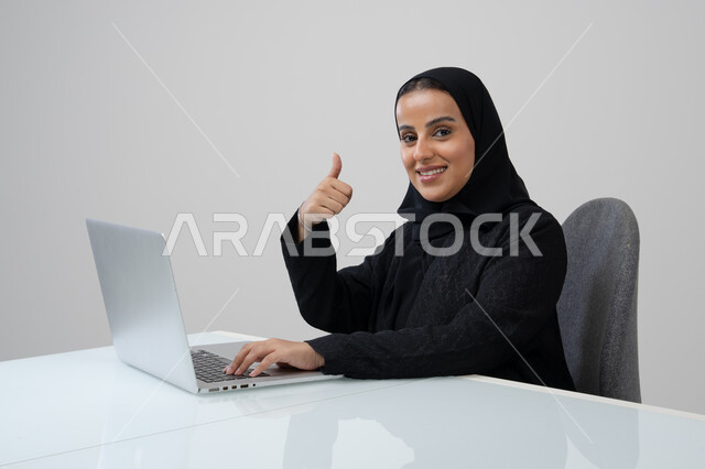 رفع اصبع الابهام بايماءات الرضى، تعابير الموافقة والقبول, مهنة ووظيفة مكتبية، ادارة وانجاز المهام باستخدام جهاز تقني حديث ومتطور, بورتريه لإمرأة عربية خليجية سعودية ترتدي الحجاب والعباءة السوداء تعمل على الحاسوب المحمول في مقر الشركة, خلفية رمادية