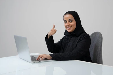 Thumbs up gesture, expressions of approval and acceptance, office profession and job, managing and completing tasks using a modern and advanced technical device, portrait of a Saudi Arabian Gulf woman wearing a hijab and black abaya working on a laptop at the company headquarters, gray background