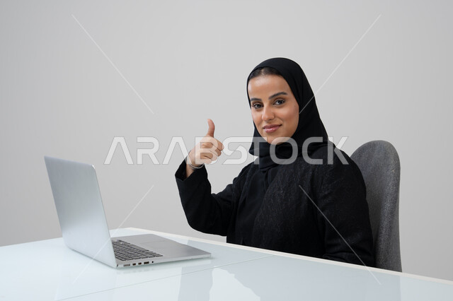 رفع اصبع الابهام بايماءات الرضى، تعابير الموافقة والقبول, مهنة ووظيفة مكتبية، ادارة وانجاز المهام باستخدام جهاز تقني حديث ومتطور, بورتريه لإمرأة عربية خليجية سعودية ترتدي الحجاب والعباءة السوداء تعمل على الحاسوب المحمول في مقر الشركة, خلفية رمادية