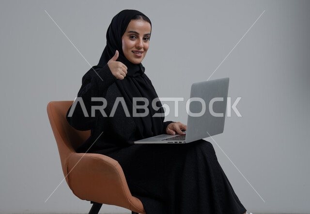 ادارة وانجاز المهام باستخدام جهاز تقني حديث ومتطور, رفع اصبع الابهام بايماءات الرضى، تعابير الموافقة والقبول, مهنة ووظيفة مكتبية، بورتريه لإمرأة عربية خليجية سعودية ترتدي الحجاب والعباءة السوداء تجلس على الكرسي وتعمل على الحاسوب المحمول, خلفية رمادية