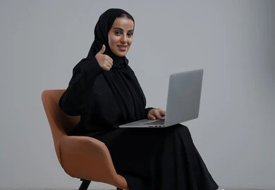 ادارة وانجاز المهام باستخدام جهاز تقني حديث ومتطور, رفع اصبع الابهام بايماءات الرضى، تعابير الموافقة والقبول, مهنة ووظيفة مكتبية، بورتريه لإمرأة عربية خليجية سعودية ترتدي الحجاب والعباءة السوداء تجلس على الكرسي وتعمل على الحاسوب المحمول, خلفية رمادية