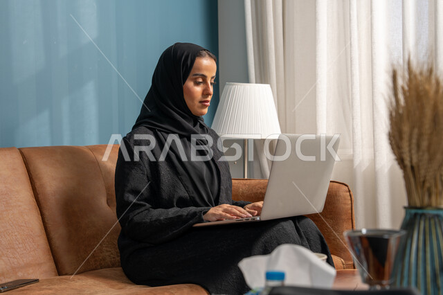 العمل على الحاسوب المحمول في صالة المنزل، ادارة وانجاز المهام باستخدام جهاز تقني حديث ومتطور, مهنة ووظيفة مكتبية، إمرأة عربية خليجية سعودية ترتدي الحجاب والعباءة السوداء تجلس على الكنبة