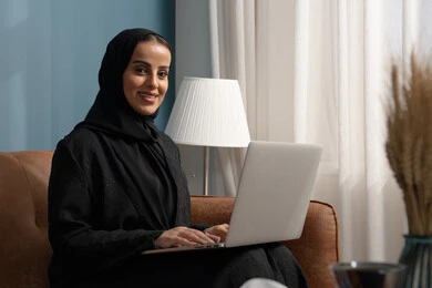 Working on a laptop in the living room, managing and completing tasks using a modern and advanced technical device, an office profession and job, a Saudi Arabian Gulf woman wearing a hijab and a black abaya sitting on the sofa
