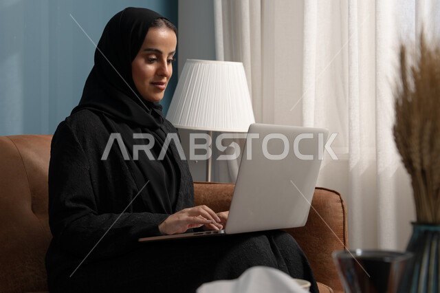 Working on a laptop in the living room, managing and completing tasks using a modern and advanced technical device, an office profession and job, a Saudi Arabian Gulf woman wearing a hijab and a black abaya sitting on the sofa