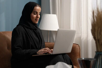 العمل على الحاسوب المحمول في صالة المنزل، ادارة وانجاز المهام باستخدام جهاز تقني حديث ومتطور, مهنة ووظيفة مكتبية، إمرأة عربية خليجية سعودية ترتدي الحجاب والعباءة السوداء تجلس على الكنبة